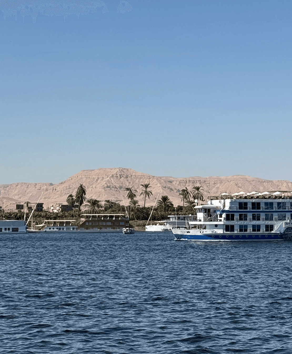 Nile River cruise ship sailing past Luxor West Bank with palm trees and mountains