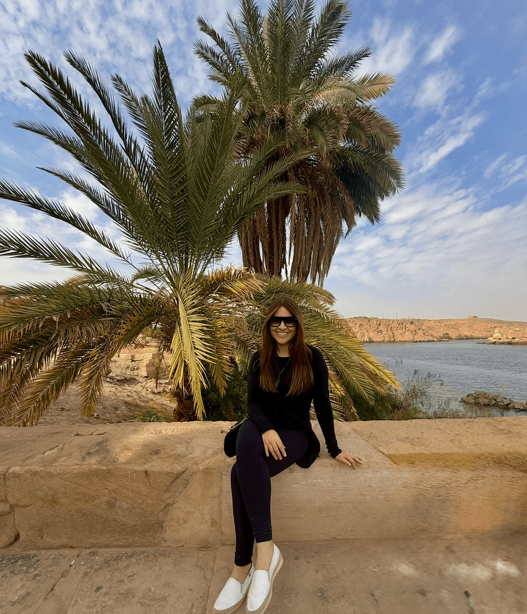 Traveler sitting by palm trees at Philae Temple with the Nile River in the background during a cruise stop